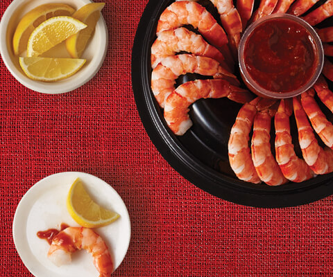 A pack shot of an open Compliments Shrimp Ring with Cocktail Sauce, a plate of lemon wedges and a small plate with a single shrimp and slice of lemon on top. 
