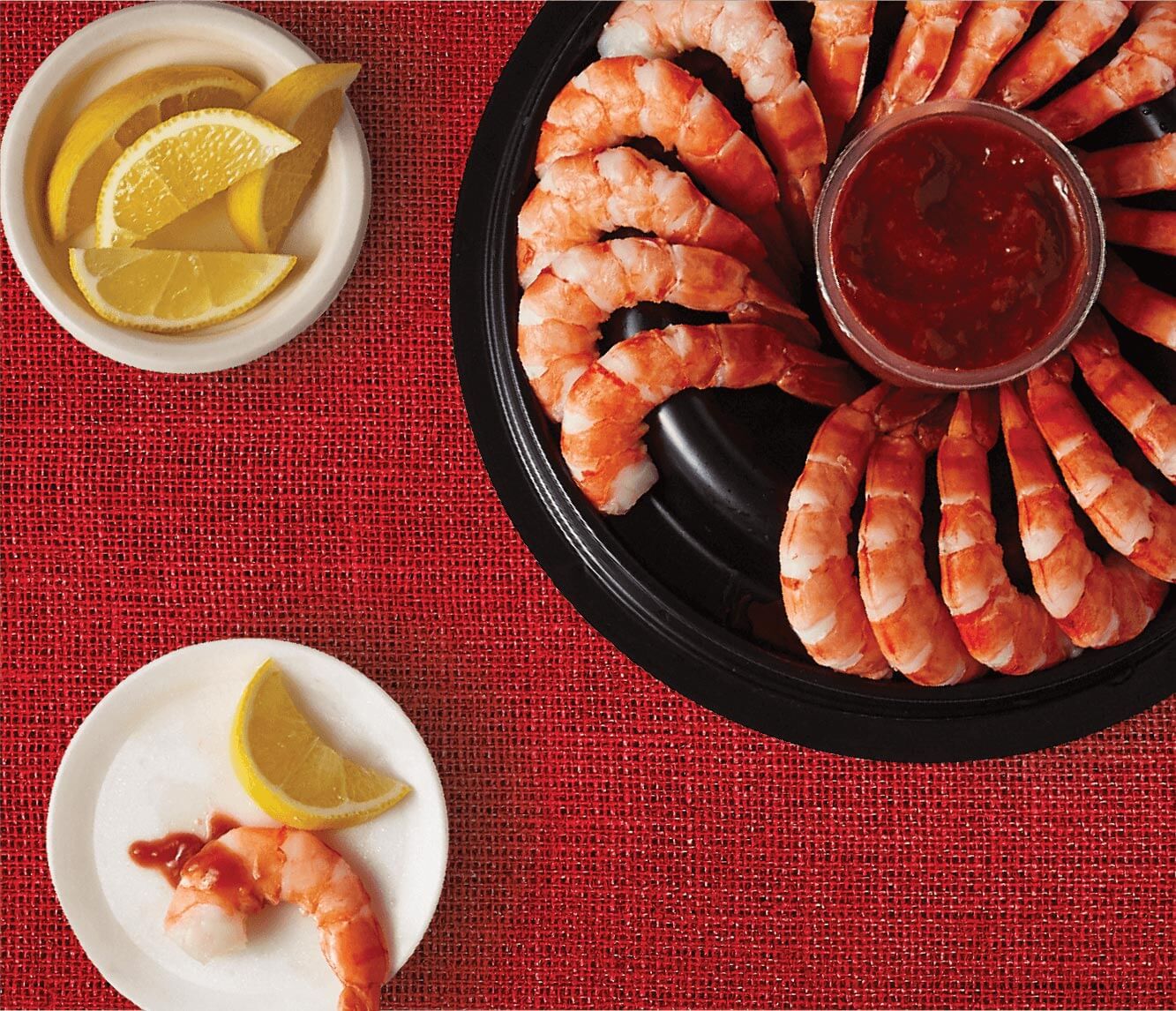 A pack shot of an open Compliments Shrimp Ring with Cocktail Sauce, a plate of lemon wedges and a small plate with a single shrimp and slice of lemon on top.