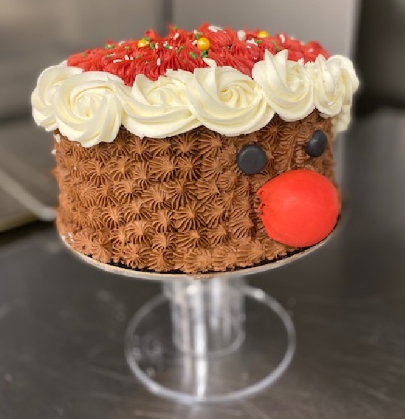 A round cake decorated with red, white and brown icing wiht a face of a reindeer with a red nose on the side.