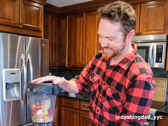 Matt stands in his kitchen blending fruits into a smoothie. A cutting board of fruits in front of him on counter